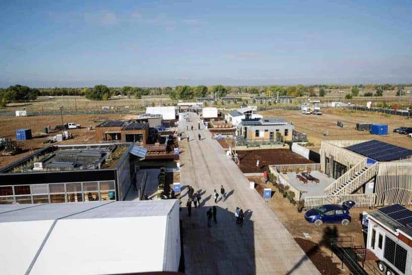 Aerial view of a long, straight road cutting through a dry flat landscape with sparse vegetation and several white-roofed buildings on either side.